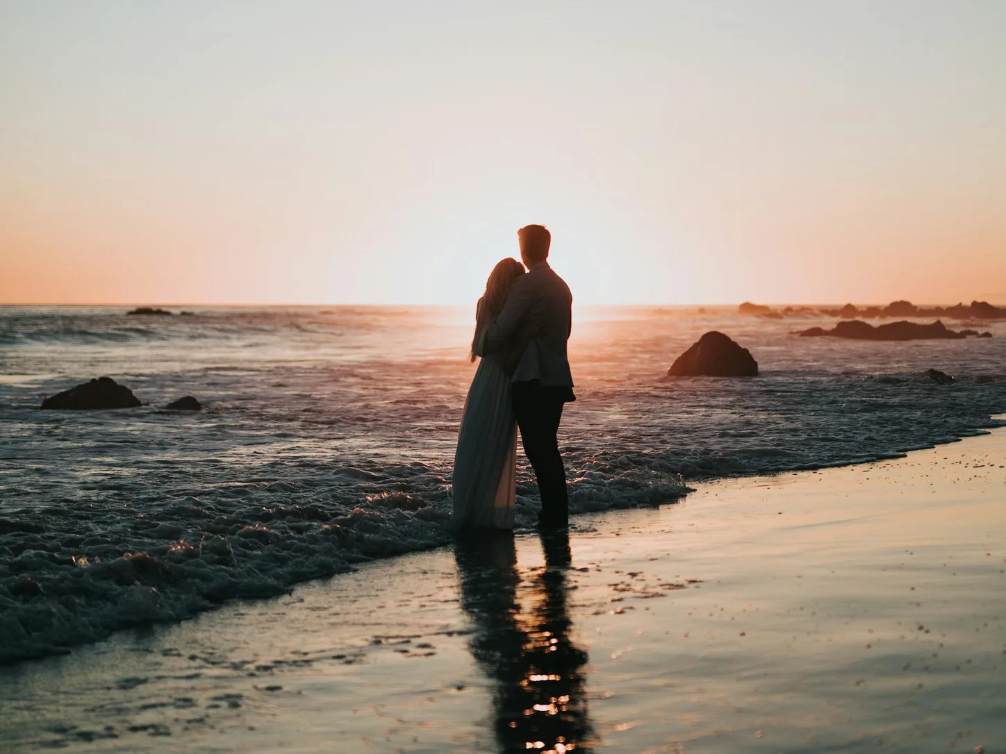 Ein Paar steht eng umschlungen im flachen Wasser am Strand, gegen die tief stehende Sonne fotografiert; die Silhouetten spiegeln sich im nassen Sand
